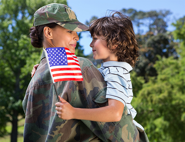 Woman in military camouflage uniform holds a young boy with an American flag in his hand