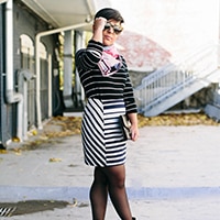 A woman with short brown hear wearing a black and white striped dress, pink scarf, black booties, round sunglasses, and has a black clutch while standing outside.