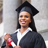 African American woman in a black graduation cap and gown with a rolled up diploma in her hand standing in front of a building with large pillars