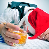 A man in a Santa hat and tie sits face down on a desk with graphs and papers on it with a glass of whiskey in his hand